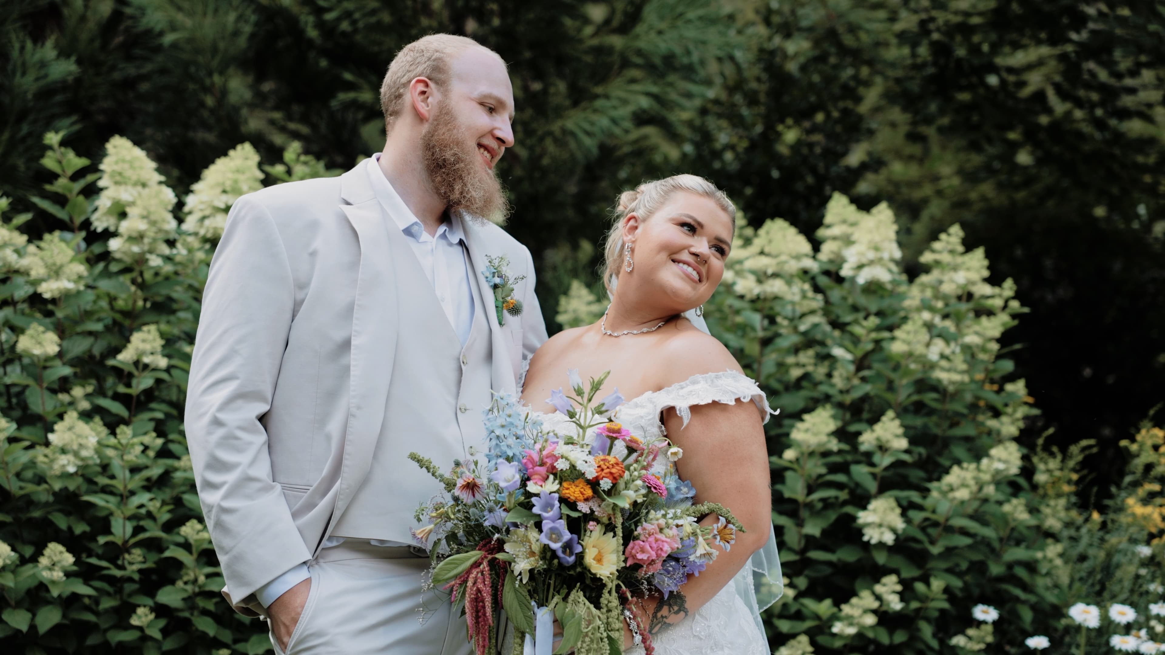 Bride holding a colorful wildflower bouquet beside her groom in a garden of white hydrangeas