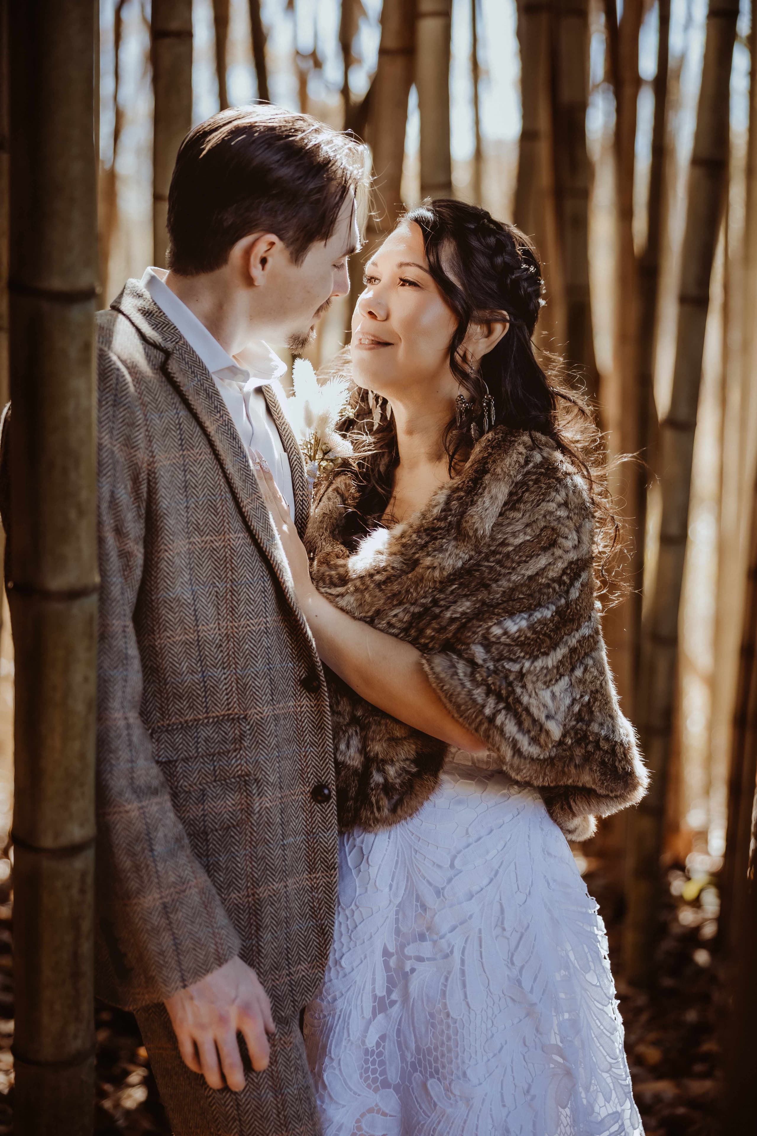 Bride in a fur stole and lace gown gazing up at the groom in a tweed suit in a sunlit bamboo grove