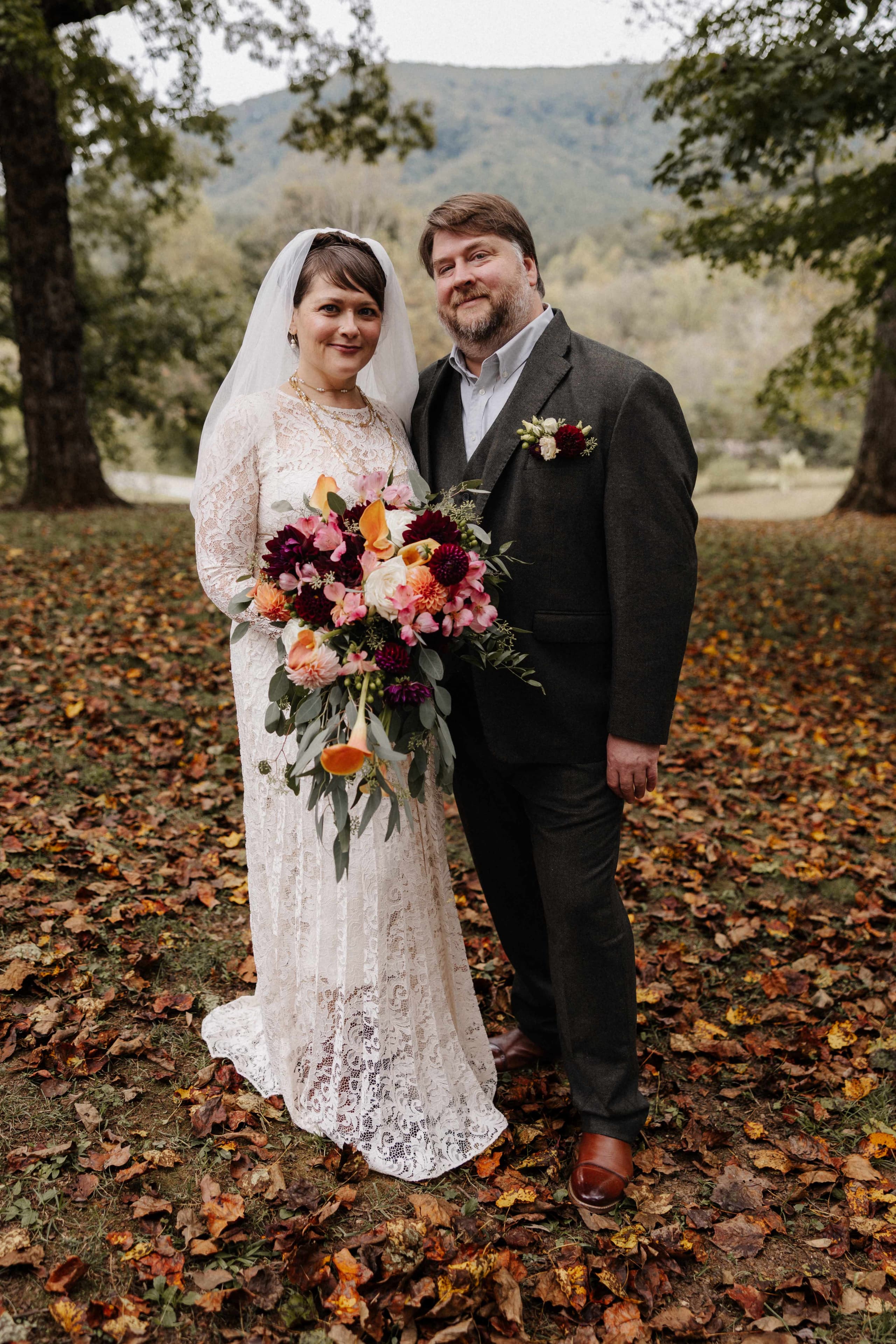 Bride in long-sleeve lace gown and groom in tweed suit standing in fall leaves with mountains behind