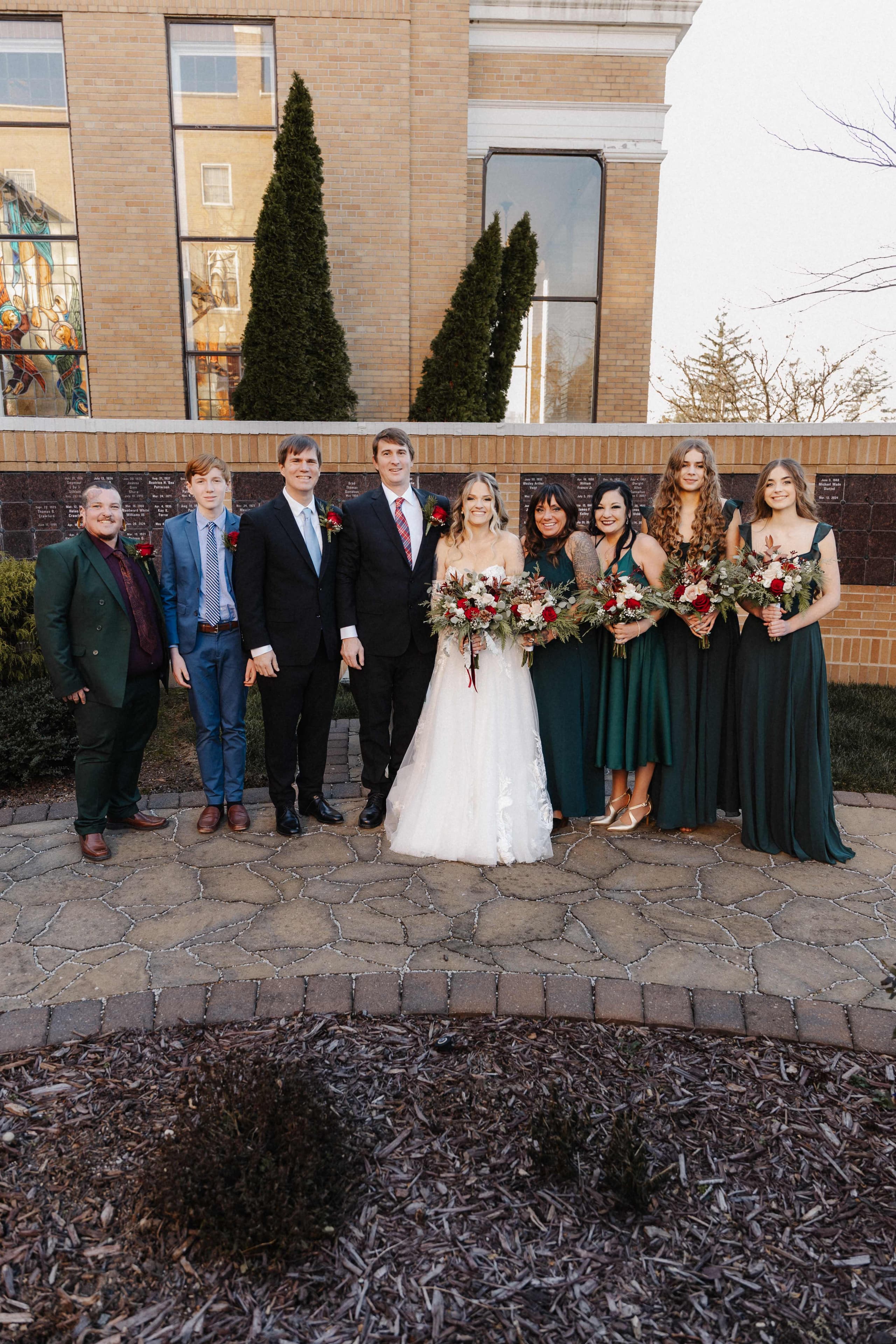 Full wedding party outside brick church with bridesmaids in emerald dresses