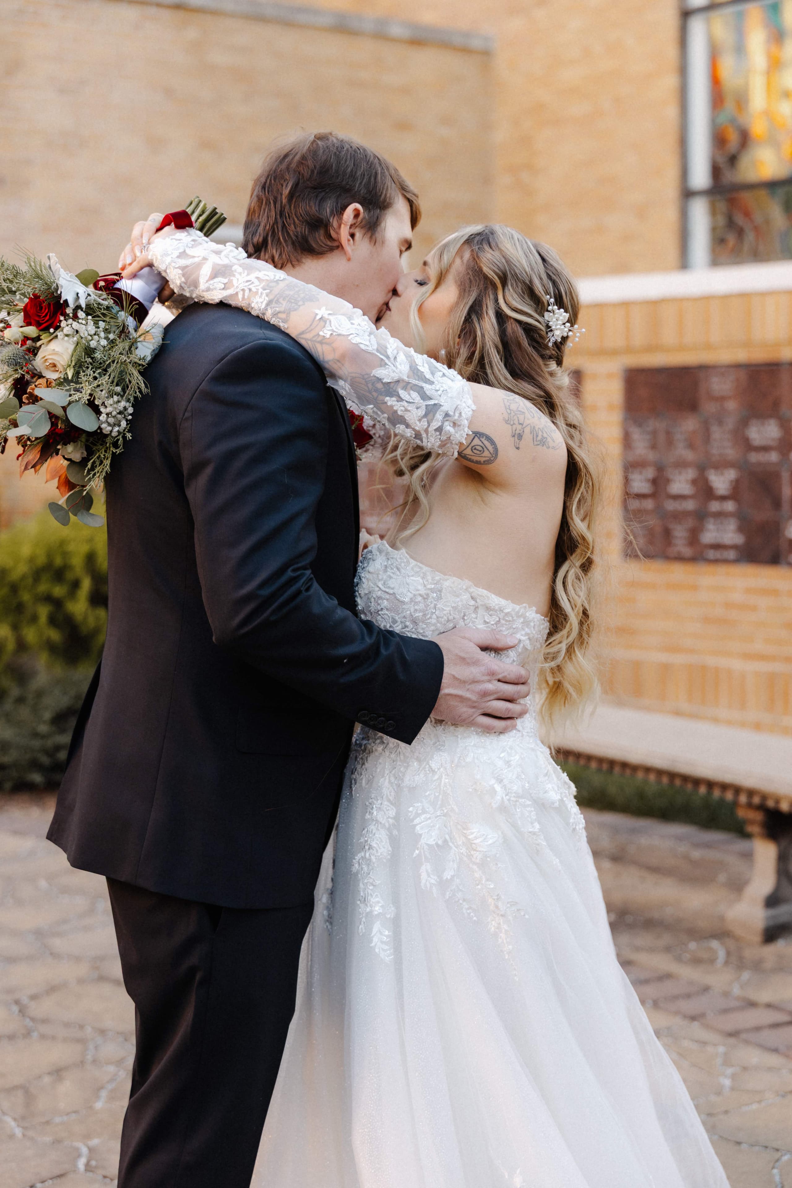 Couple kissing outside church with winter bouquet showing bride's back tattoos
