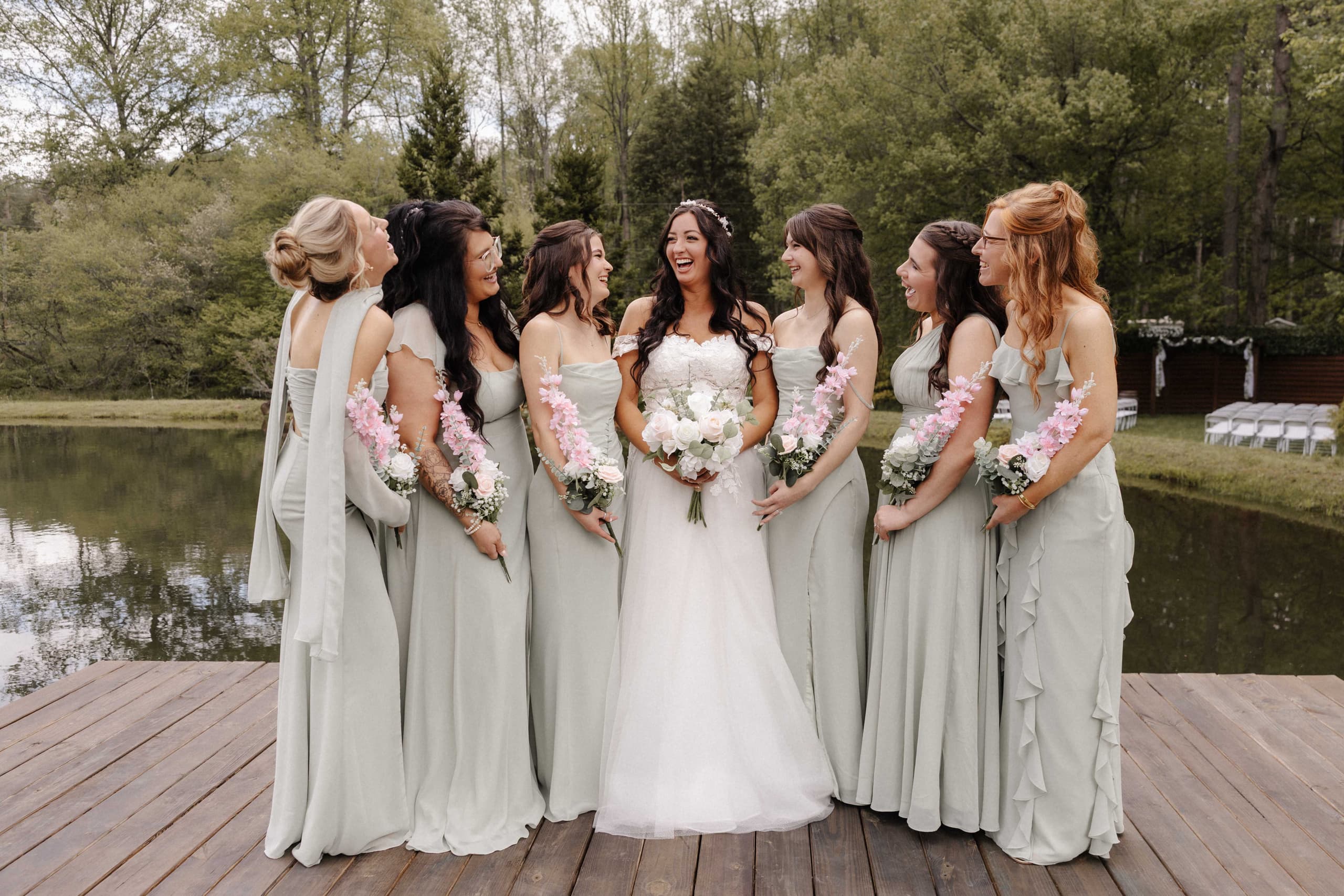 Bride laughing with bridesmaids in sage green dresses on dock by lake