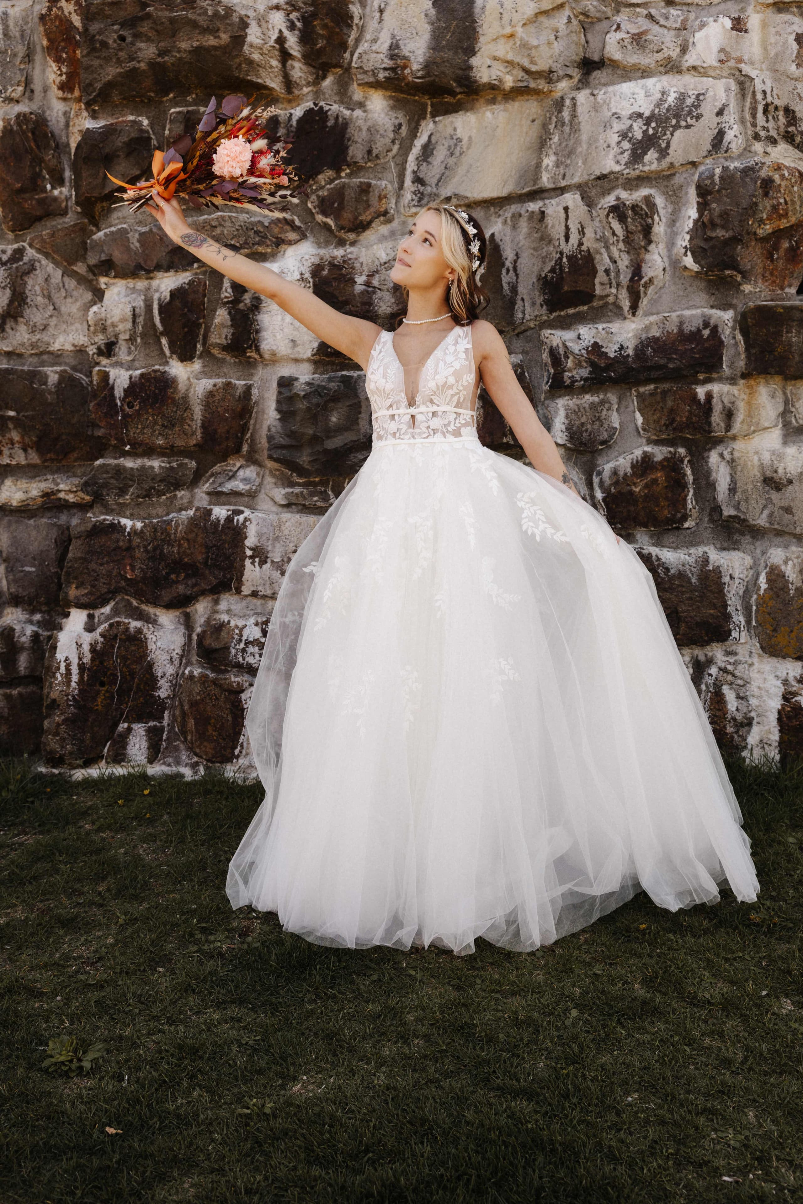 Bride raising bouquet by stone wall in ballgown dress
