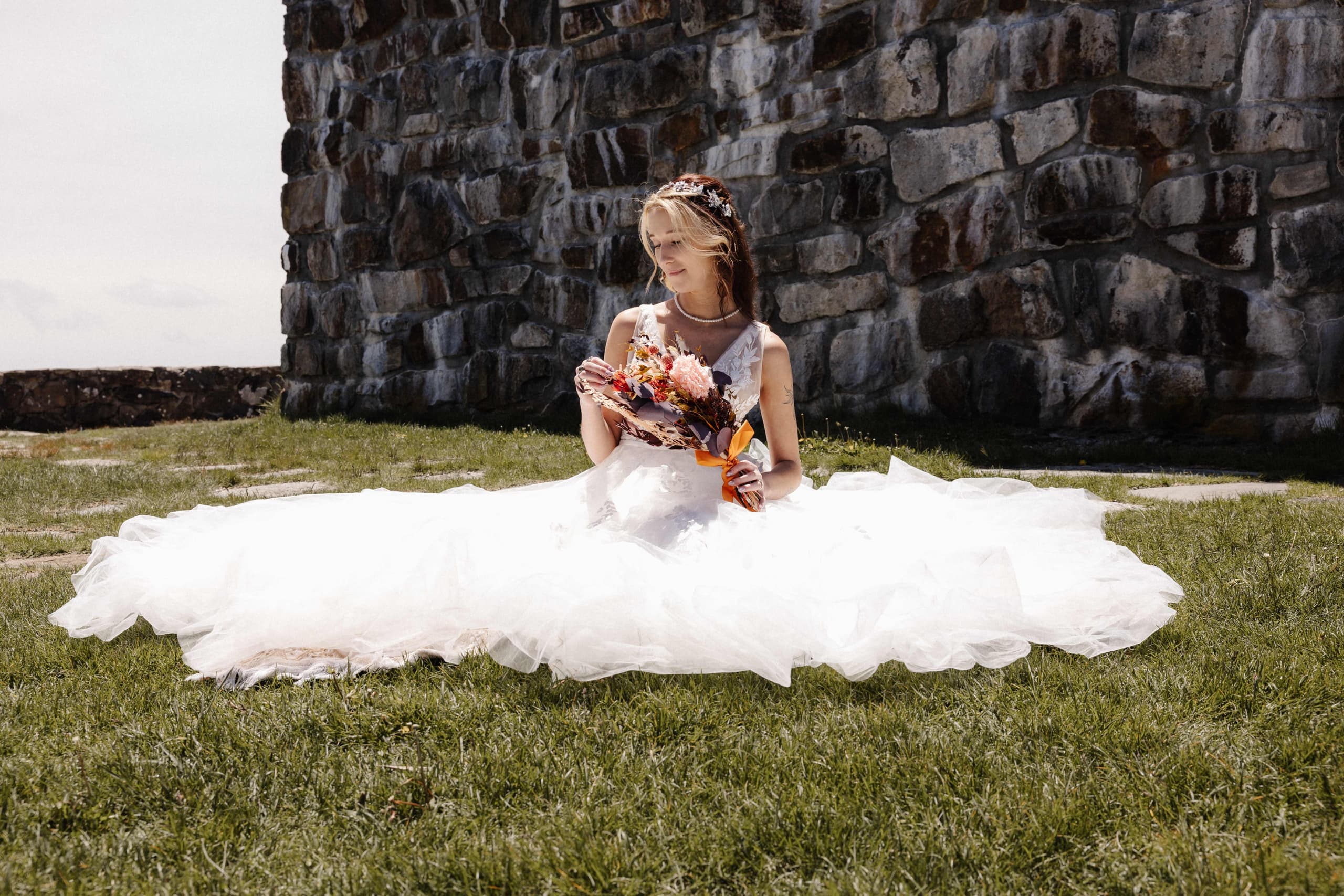 Bride sitting with dress spread on grass by stone wall