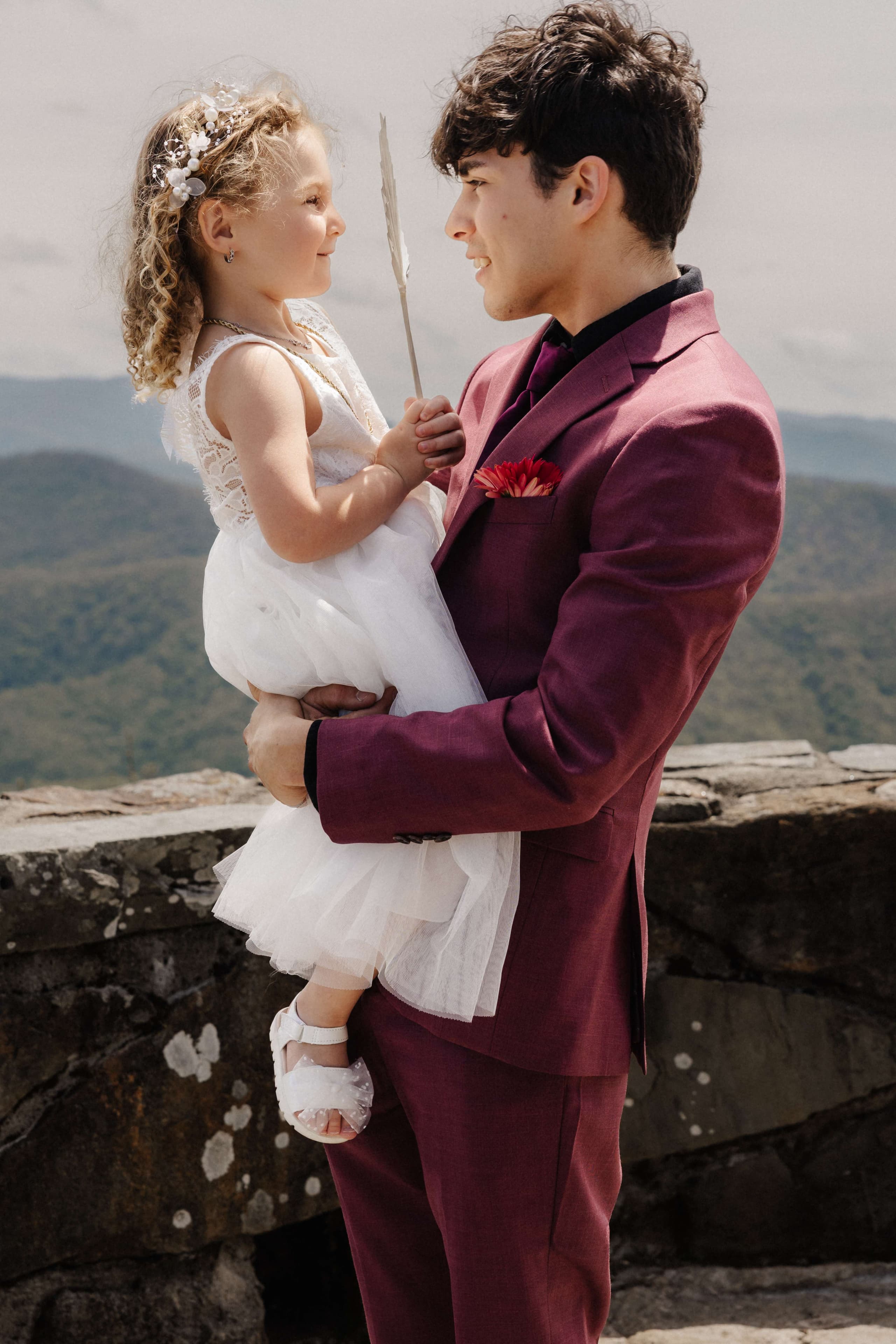 Groom in burgundy suit holding flower girl with mountain backdrop